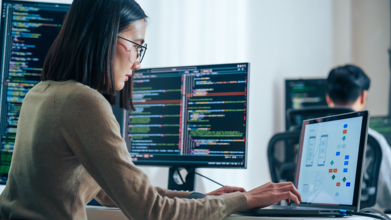 Woman reviewing three monitors with code and process flow